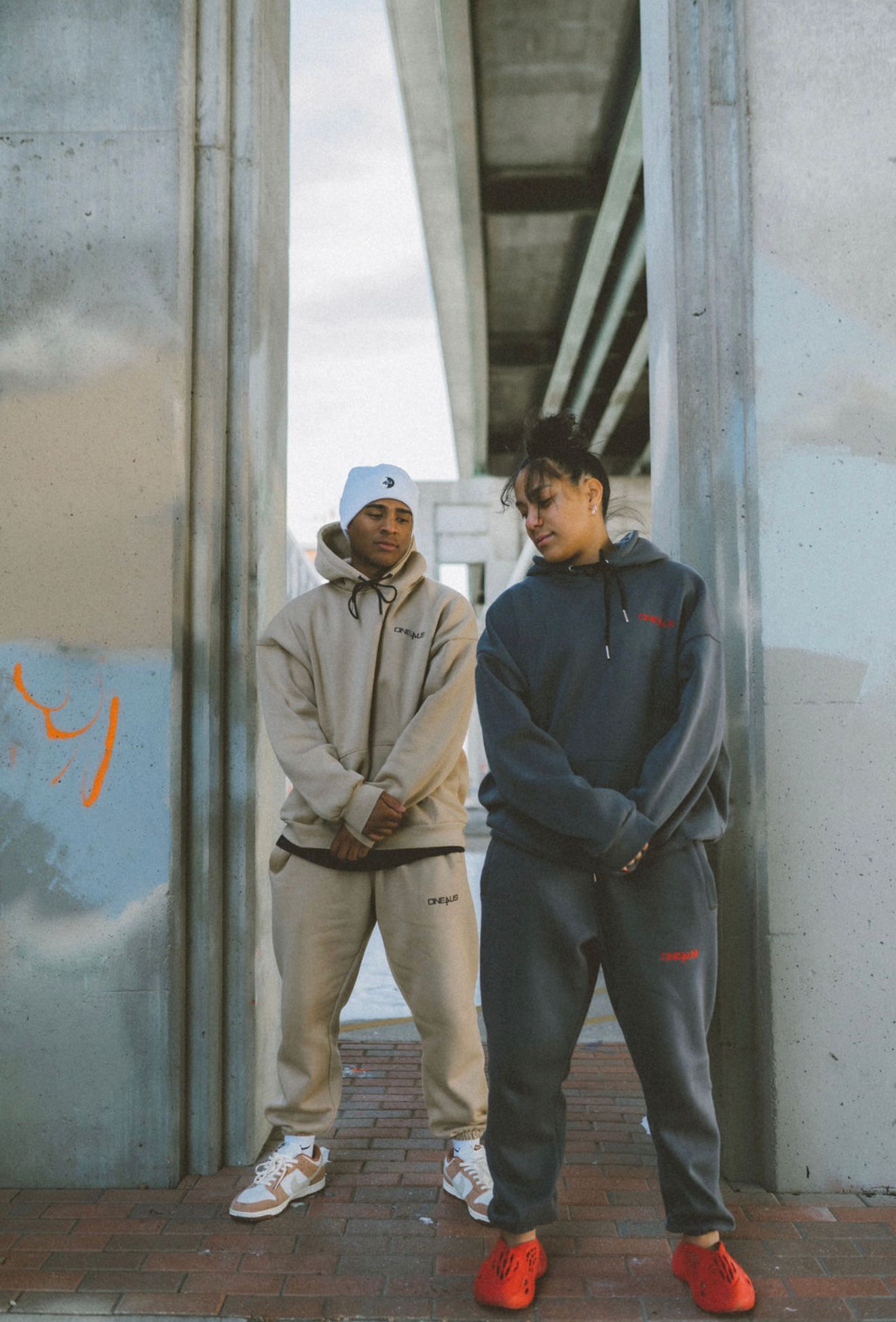 Man and woman wearing tracksuits standing against a concrete wall with graffiti.